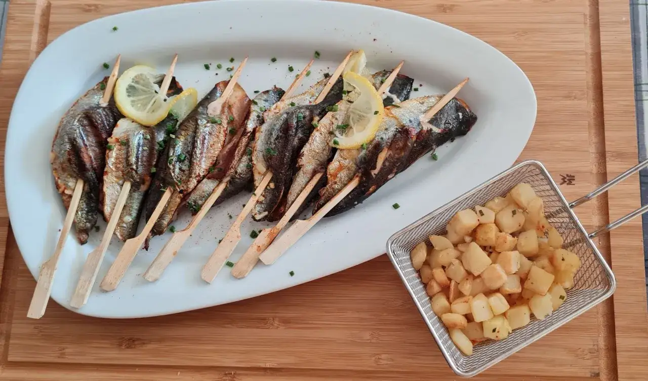 Sardines grillées sur brochettes, garnies de citron et persil, accompagnées de pommes de terre sautées dans un panier métallique.